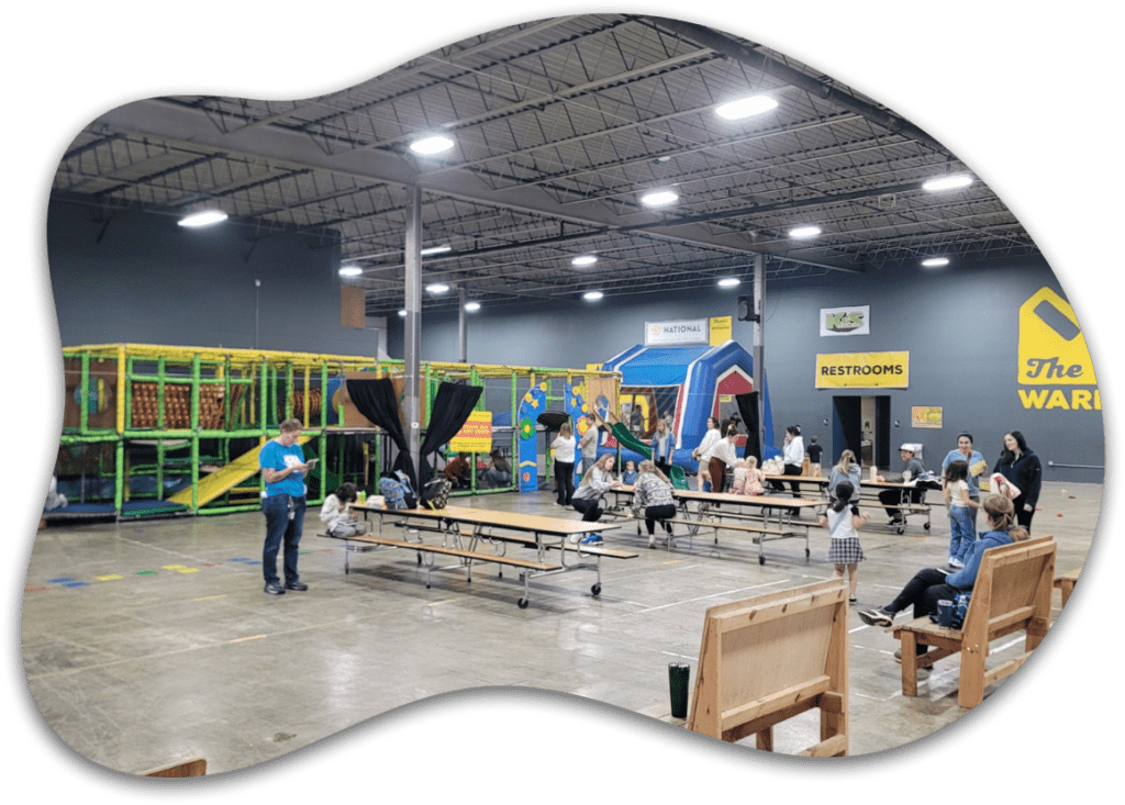 Wide view of the Kidzone play area at The Warehouse featuring a jungle gym structure, inflatable bounce house, picnic tables, and families inside an organic-framed graphic layout.