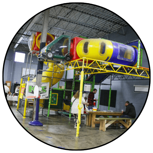 Circular crop photo of the Kidzone indoor play area at The Warehouse, featuring a colorful multi-level play structure with tunnels and slides, a mini basketball hoop, and seating tables.