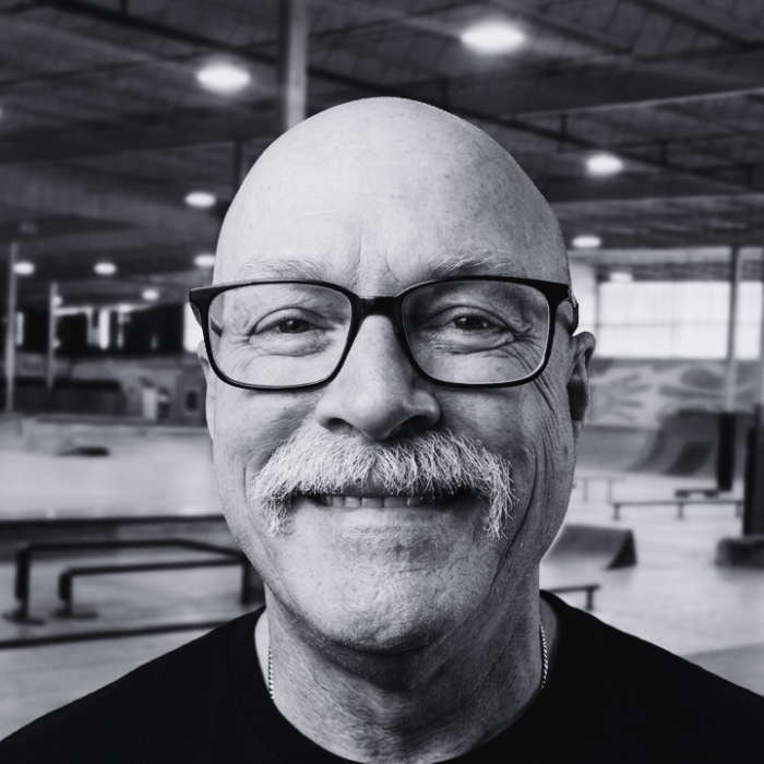 Headshot of Dr. Perry Griffeth, Vice President of the Board of Directors at The Warehouse, smiling broadly with a gray chevron mustache and black-rimmed glasses inside an indoor skatepark facility.