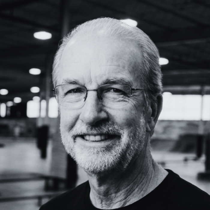 Headshot of Curtis Schopp, Board Member at The Warehouse, smiling with a short gray beard and thin-rimmed glasses inside an indoor skatepark facility.