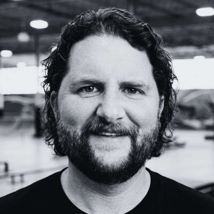Headshot of Joe Sheese, Board Member at The Warehouse, smiling with wavy dark hair and a full beard inside an indoor skatepark facility.