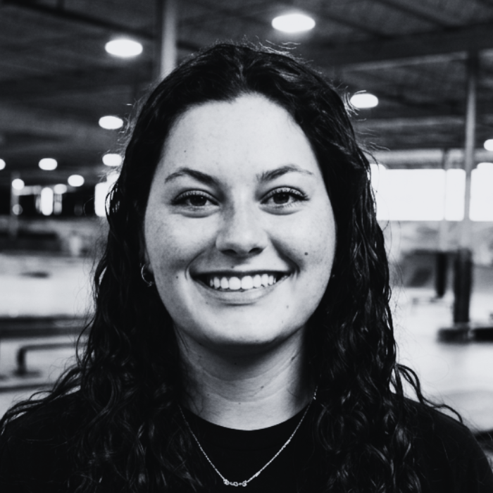 Headshot of Lexi, Floor Staff member at The Warehouse, smiling warmly with long curly dark hair and a delicate necklace inside an indoor skatepark facility.