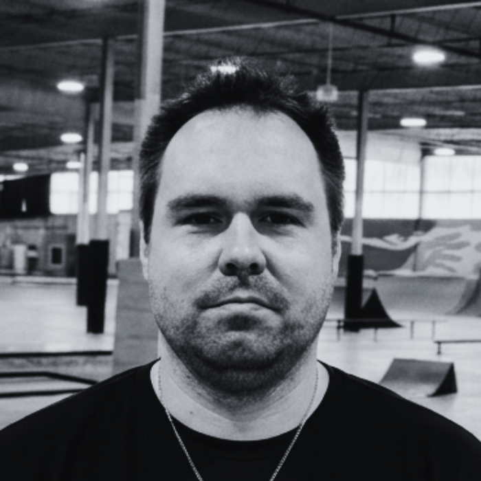 Headshot of Nick, Certified Trainer and Floor Staff at The Warehouse, wearing a dark shirt and a silver cross necklace inside an indoor skatepark facility.