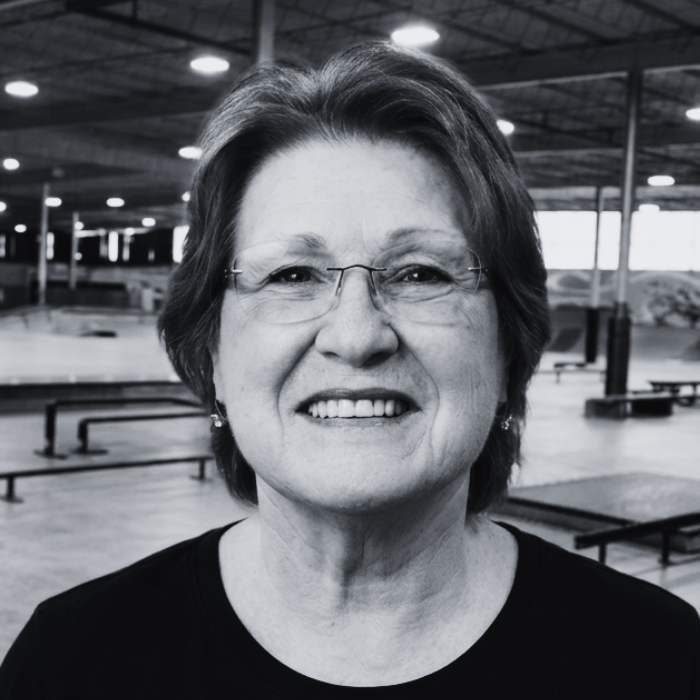 Headshot of Rhonda, Director of Kids Programming at The Warehouse, smiling warmly while wearing rimless glasses inside an indoor skatepark facility.