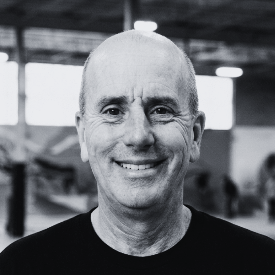 Staff portrait of David Weil, Executive Director of The Warehouse. He is a man with short brown hair and glasses, smiling warmly while wearing a dark zip-up jacket. He is standing inside the indoor sports facility at The Warehouse in Bloomington, Indiana, with basketball hoops, a concrete floor, and natural light streaming in from large windows blurred in the background.
