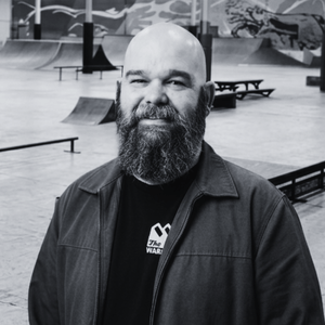 A monochrome medium portrait of Josh Utterback, a bald man with a grayscale graying beard, smiling while standing in the black and white interior of The Warehouse Bloomington's indoor skatepark. He wears a dark jacket over a black t-shirt with a monochrome Warehouse logo. Grayscale ramps and rails are in the background.