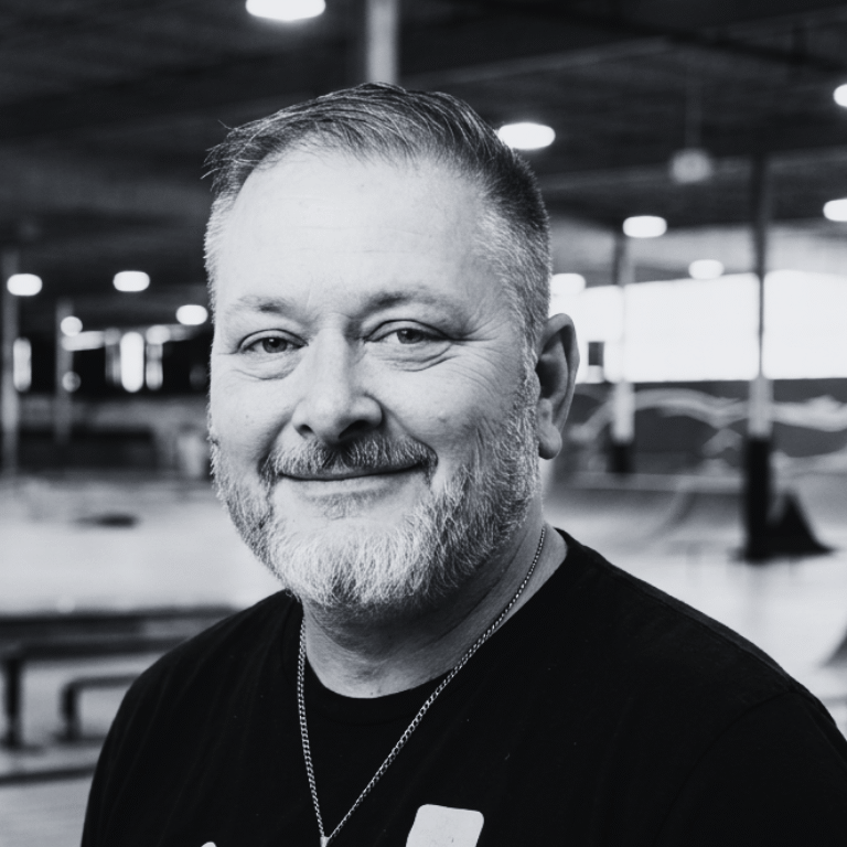 Staff portrait of Spencer Armstrong, a young man with short brown hair and a light beard, smiling while wearing black t-shirt. He is standing inside the indoor recreation space at The Warehouse in Bloomington, Indiana, with the indoor skate park behind him, and an open warehouse ceiling blurred in the background.
